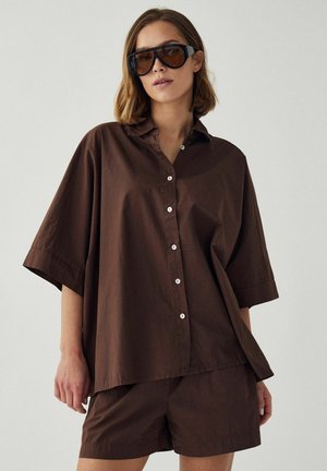 Kvinde iført oversize brun skjorte med knapper og matchende shorts, store mørke solbriller, stående mod en ensfarvet baggrund.