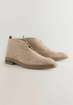 Botas de tobillo de ante beige con una textura suave, puntera redondeada y diseño de cordones. Cuentan con un tacón bajo y una costura en contraste a lo largo de la suela.