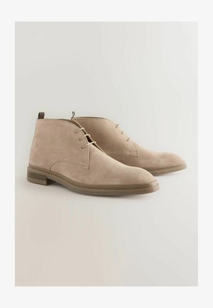 Bežové semišové členkové topánky s hladkou textúrou, zaoblenou špicou a šnurovacím dizajnom. Obsahujú nízky opätok a kontrastné šitie pozdĺž podrážky.