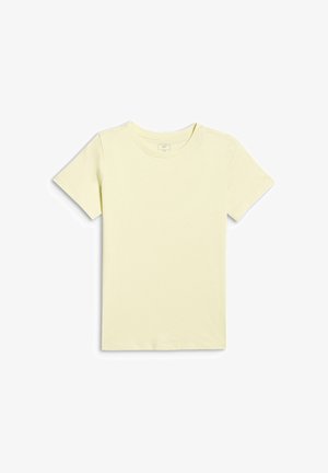 Jeune fille debout, vêtue d'un t-shirt jaune pâle, d'un pantalon évasé gris, de baskets blanches, tenant un sweat rose dans sa main gauche.