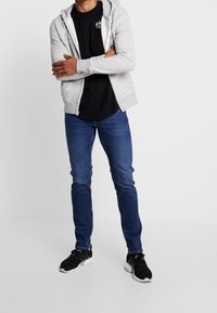 Felpa grigia con zip sopra una t-shirt nera, jeans blu e scarpe sportive nere. Tessuto liscio e design aderente con un aspetto semplice.