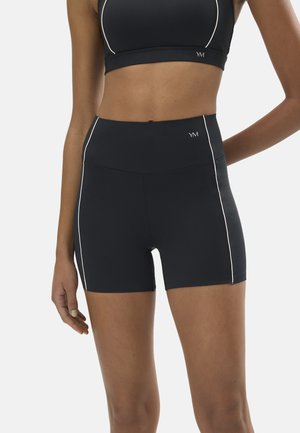 Mujer usando pantalones cortos deportivos de talle alto negros y un sujetador deportivo a juego con ribete blanco y logo "YM" en los pantalones cortos y el sujetador.