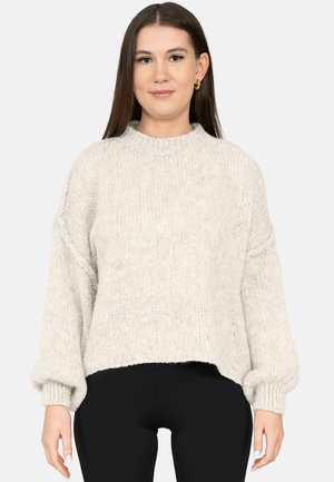 Vrouw met recht bruin haar, goudkleurige ronde oorbellen, een lichtgrijze gebreide trui met ballonmouwen en een zwarte broek, die recht vooruit kijkt.