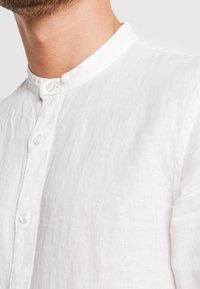 Camicia bianca di lino con colletto alto, dotata di piccoli bottoni lungo il davanti e un tessuto testurizzato che si adagia dolcemente.
