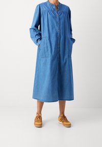 Denimkjole i lyseblå med lange ærmer, knaplukning foran, rund halsudskæring og to sidelommer, parret med brune sneakers.