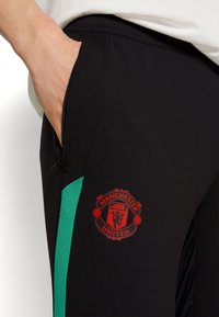 Černé sportovní kalhoty s zeleným bočním panelem a červeným logem Manchester United na stehně. Obsahují kapsu na zip a mají hladkou texturu.