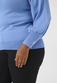 Jasnoniebieski sweter dzianinowy z bufiastymi rękawami i akcentami w postaci guzików przy mankietach, w połączeniu z ciemnymi czarnymi spodniami. Gładka tekstura i dopasowany fason.