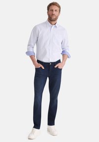 Helles, blaues Button-Down-Hemd mit kleinen dunklen Punkten, marineblaue Jeans und weiße Sneakers. Das Hemd hat eine linke Brusttasche und aufgerollte Ärmel.