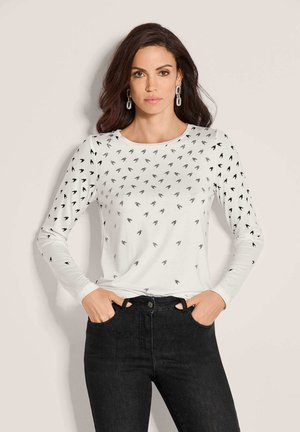 Vrouw met lang donker haar die zilveren kettingoorbellen draagt, een wit shirt met lange mouwen met zwarte vogelprints en een zwarte broek, staand met handen in de zakken.