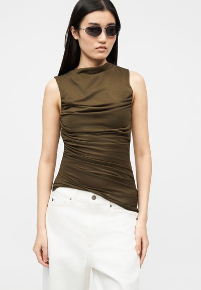 Sötét olívazöld redőzött ujjatlan felsőt, fehér nadrágot és fekete ovális napszemüveget viselő nő áll sima világos háttér előtt.