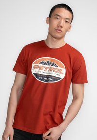 Camiseta de algodón roja con un estampado gráfico que presenta una silueta de montaña y el texto "PETROL". Manga corta, cuello redondo y corte relajado.