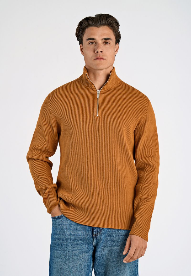 Maglione a coste color senape con colletto alto e mezza zip, abbinato a jeans in denim blu, in piedi su uno sfondo chiaro.