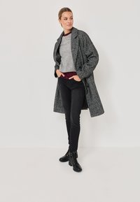 Abrigo gris con textura y solapas grandes, llevado sobre una blusa gris claro y jeans ajustados negros. Botines negros con cordones completan el atuendo.