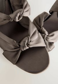 Flache Sandalen mit geknoteten Stoffriemen. Hergestellt aus weichem, mattem, taupefarbenem Material. Rundliche Zehen und glatte, flache Sohlen. Einfaches und zeitgemäßes Design.