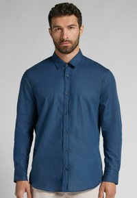 Långärmad, knappad skjorta i mörkblått tyg. Har en klassisk krage, framknappar och rak nederkant. Strukturerat, lätt material.