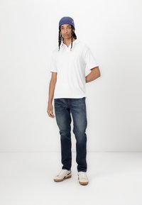 Camisa polo blanca de manga corta con rayas, gorro de punto azul, jeans oscuros y zapatillas blancas con acentos beige, de pie frente a un fondo blanco.
