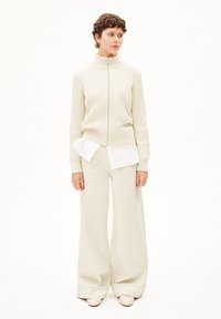 Beige Strickjacke mit hohem Kragen, die über einem weißen Hemd und einer weit geschnittenen, hellbeigen Hose getragen wird, abgerundet mit elfenbeinfarbenen Schuhen.