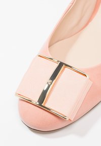 Ballerine en daim rose avec un grand nœud en tissu rose et un accent en métal doré sur l'avant.