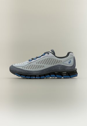 Hellgrauer Sportschuh aus Mesh- und Wildledermaterialien. Verfügt über eine schwarz-blaue, gepolsterte Sohle, horizontale Ausschnitte und graue Schnürsenkel.