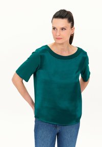 Teal satijnen blouse met korte mouwen, brede halslijn met plooien, gladde textuur en een relaxte pasvorm, gedragen met blauwe jeans.