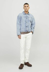Giacca di jeans azzurro chiaro con tasche frontali, maglia a girocollo marrone sotto, pantaloni bianchi a gamba dritta e scarpe marroni con lacci.
