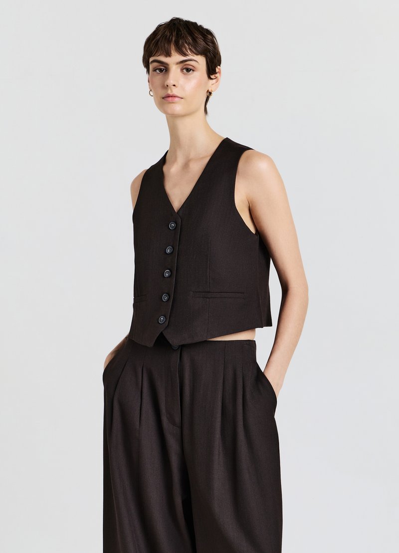 Schwarze ärmellose Weste mit V-Ausschnitt, ausgestattet mit sechs schwarzen Knöpfen und Seitentaschen, kombiniert mit hoch taillierten, weit geschnittenen Hosen.