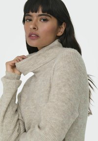 Pull à col roulé beige avec un motif de tricot texturé et un col replié. Manches longues avec des poignets côtelés, tissu doux, coupe décontractée.