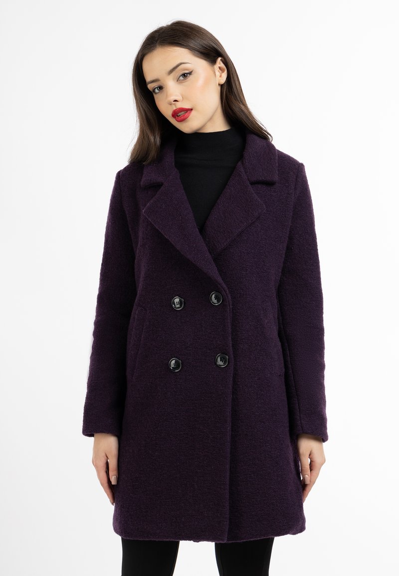 Dubbelknäppt kappa i djup lila tyg, med en texturerad yta, snedskurna slag och svarta knappar. Modellen bär en svart polotröja.