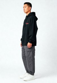 Czarna bluza z kapturem z kieszenią kangurką oraz czerwonym napisem, zestawiona z szarymi spodniami cargo i białymi sneakersami. Materiał wydaje się miękki i swobodny.