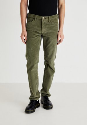 Person wearing olive green corduroy pants with black leather lace-up shoes standing against a plain white background.