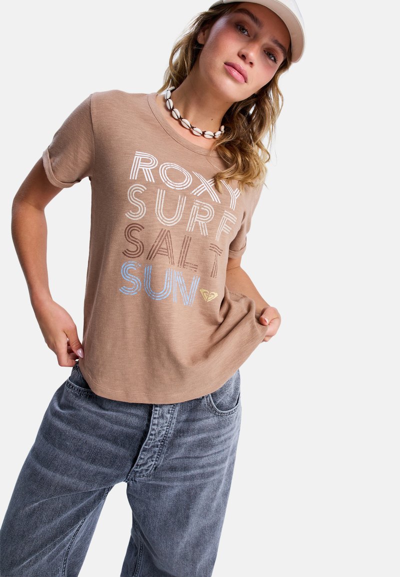 Jonge vrouw die een beige pet, een schelpenketting en een bruin T-shirt met de tekst "ROXY SURF SALT SUN" draagt, in combinatie met losse grijze jeans.