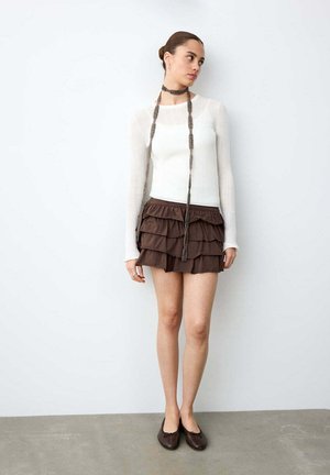 Jeune femme debout contre un mur blanc, portant un haut blanc à manches longues, une mini-jupe marron à volants superposés, un long collier et des ballerines marron.