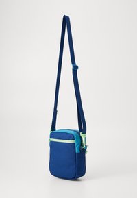 Saco cruzado azul com forma retangular, feito de tecido. Apresenta um topo em turquesa contrastante e detalhes em verde no fecho.