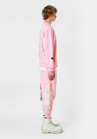 Sudadera rosa con gráficos de alas, combinada con pantalones de chándal a juego que presentan diseños similares. Con botas de combate blancas.
