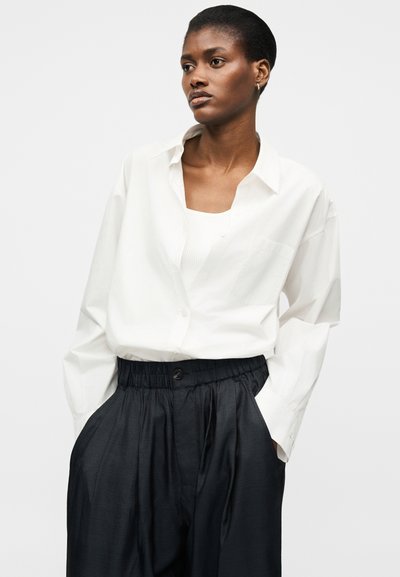 Γυναίκα που φοράει χαλαρό λευκό πουκάμισο με κουμπιά πάνω από λευκή μπλούζα και ψηλόμεσο παντελόνι με μαύρα, φαρδιά πόδια, με τα χέρια στις τσέπες.