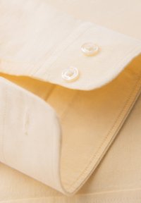 Poignet de chemise beige avec deux boutons transparents cousus, montrant une couture détaillée et la texture du tissu.