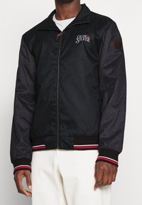 Giacca nera con maniche grigio scuro a contrasto, chiusura con zip, polsini a coste con strisce rosse e bianche, e logo Quiksilver sul petto.