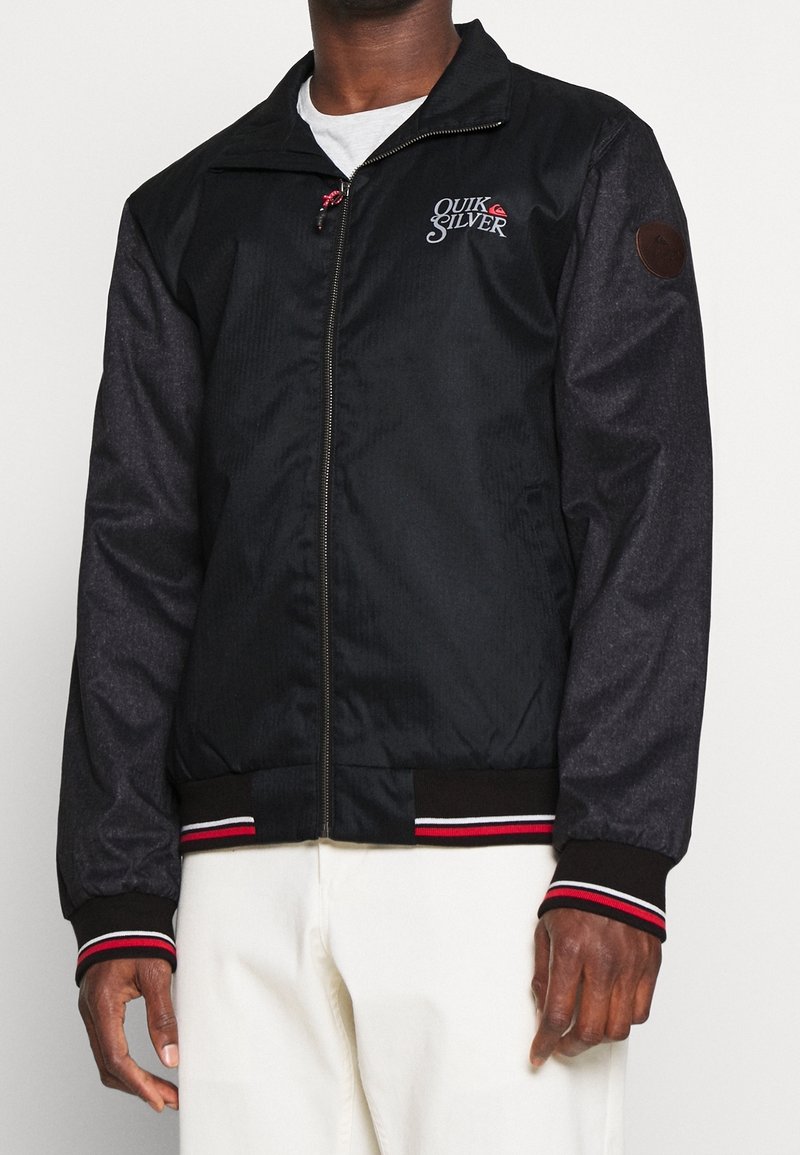 Giacca nera con maniche grigio scuro a contrasto, chiusura con zip, polsini a coste con strisce rosse e bianche, e logo Quiksilver sul petto.