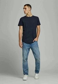 Jovem em pé, a usar uma t-shirt preta, jeans azul claro e ténis brancos, com uma mão no bolso, a olhar para a esquerda.