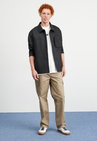 Chemise noire à boutons avec poche poitrine, portée sur un t-shirt blanc. Pantalons cargo beiges avec poches latérales. Baskets blanches avec accents navy.
