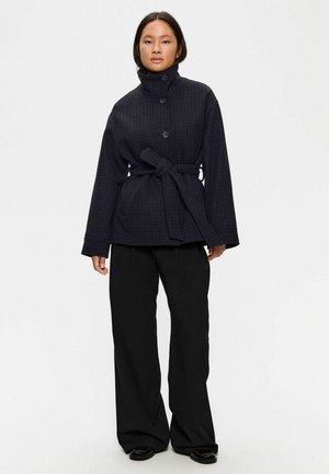 Mujer con chaqueta oscura texturizada con cinturón, grandes botones y cuello alto, combinada con pantalones negros holgados y zapatos negros.