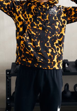 Person iført en gul og sort leopardprint Nike-hættetrøje og sorte Nike-bukser står foran træningsvægte og kettlebells.