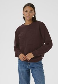 Sweatshirt bordeaux avec le numéro en relief « 27 » �à l'avant, col rond et poignets côtelés ; associé à un jean en denim bleu.