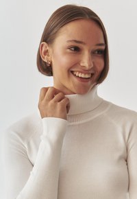 Maglione bianco a collo alto con collo e polsini a coste, design aderente, texture in maglia liscia, indossato da una persona con capelli corti castani.
