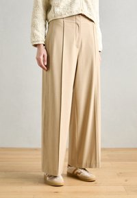 Beige, wide-leg trousers with front pleats, made from a smooth fabric, paired with cream sneakers featuring subtle beige accents.