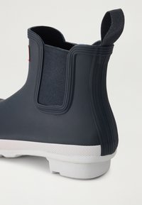 Botas de lluvia de goma negra con un panel lateral elástico texturizado, suela blanca y un pequeño lazo trasero para facilitar el uso. Presentan estrías verticales.