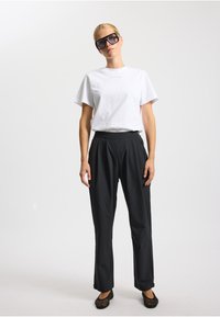 Hvid bomulds T-shirt kombineret med sorte plisserede bukser. Modellen bærer store sorte solbriller og flade sko i sort netmateriale. Ensfarvet baggrund.