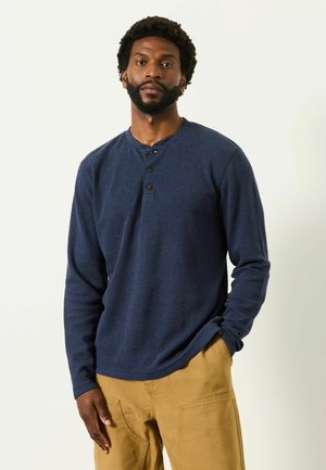 Man with beard wearing a navy blue long-sleeve henley shirt and tan pants standing with one hand in pocket against a plain background.