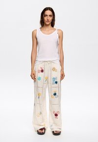 Weißes Tanktop mit rundem Ausschnitt. Lockere beige Hose mit buntem Blumen-Stickerei und Spitzenverzierung, kombiniert mit schwarzen Sandalen.