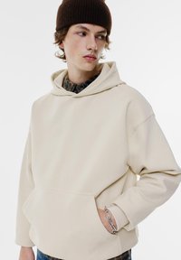 Beige huvtröja med känguruficka och ribbade muddar. Mjukt material, avslappnad passform. Buren över en rutmönstrad skjorta.
